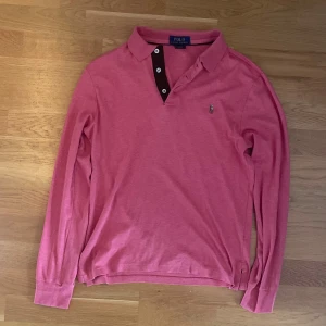 Rosa långärmad pikétröja Polo Ralph Lauren - Snygg rosa långärmad pikétröja från Polo Ralph Lauren i slim fit. Klassisk krage, knäppning med tre knappar och broderad logga på bröstet. Tillverkad i mjuk bomull som känns skön mot huden. Perfekt för dig som gillar stilrena och sportiga plagg.