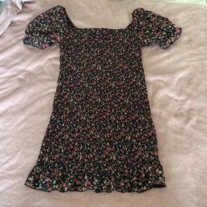 Blommig kortklänning från FB Sister - Supersöt kortklänning från FB Sister med svart botten och små blommor i rosa, rött och grönt. Klänningen har puffiga korta ärmar, smockad överdel och volangkant nertill. Perfekt för sommarens alla tillfällen!