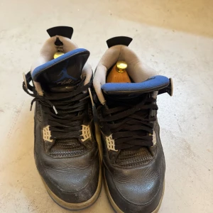 Nike Air Jordan 4 Retro svart/blå 46 - Säljer ett par Nike Air Jordan 4 Retro i svart skinn med blå och grå detaljer. Skorna är väldigt slitna och jag ber inte om mycket mer än att någon tar över dom. Priset är 350kr och storlek 46