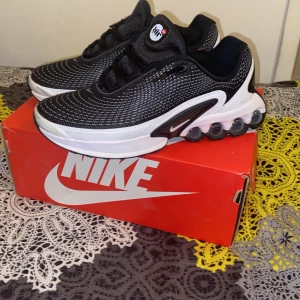 Svarta Nike Air Max Dn - Säljer ett par svarta Nike sneakers med vit och svart sula och coola luftbubblor i hälen. Ovandelen är i mesh med snyggt mönster och klassisk Nike-logga på sidan. Skorna har snörning och en sportig vibe, perfekta för dig som gillar streetstyle.
