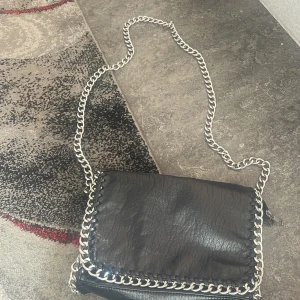Svart axelväska med kedjedetaljer - Cool svart axelväska i skinnimitation med grov silverfärgad kedja som axelrem och runt kanterna. Väskan har lock och dragkedja på baksidan, perfekt för att hålla koll på dina grejer. Snygg och edgy design som sticker ut.