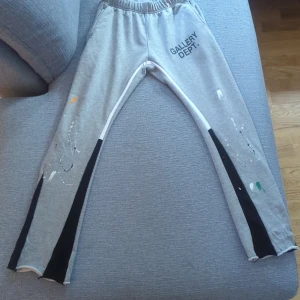 Grå mjukisbyxor Gallery Dept S - Grå mjukisbyxor från Gallery Dept. med svart och vit panel längs benen och färgstänk i vitt, grönt och orange. Byxorna har elastisk midja och är tillverkade i 100% bomull. Snygg streetstil med unik design och tryckt logga på ena låret.