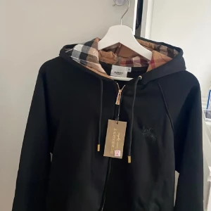 Svart Burberry hoodie med rutigt foder - Svart hoodie från Burberry med klassiskt rutigt foder i huvan. Guldiga detaljer på dragkedjan och snörningen. Stort Burberry-märke på bröstet och mjukt material som känns lyxigt. Perfekt för dig som vill ha en stilren och ikonisk look.