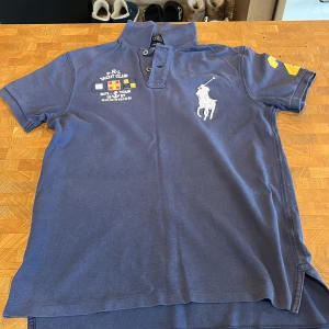 Marinblå Polo Ralph Lauren t-shirt S - Marinblå t-shirt från Polo Ralph Lauren med klassisk polokrage och två knappar. Stor vit polospelare på bröstet, broderade Yacht Club-detaljer och gul siffra 2 på ärmen. Custom fit-modell i mjuk bomull som sitter snyggt och sportigt.