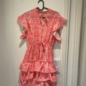 Rosa mönstrad volangklänning  - Supersöt kortklänning från Hailys i rosa och vitt med volanger och knytning i halsen. Klänningen har ett unikt mönster, korta volangärmar och resår i midjan för en snygg passform. Perfekt för sommarens alla tillfällen! Aldrig använd!!