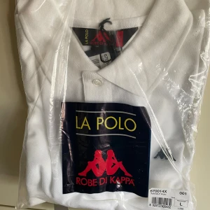 Robe di Kappa polo t-shirt vit stl L - Vit klassisk polo t-shirt från Robe di Kappa i storlek L. T-shirten har krage, korta ärmar och knapp framtill. Materialet är bomull och den har en stilren look med diskret logga på bröstet. Perfekt för dig som gillar sportig och clean stil. NOS i obruten förpackning.
