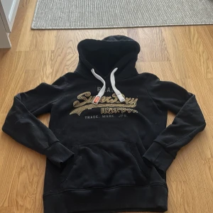 Svart hoodie från Superdry med tryck - Svart hoodie från Superdry med guldigt/glittrigt tryck och japanska tecken på bröstet. Tröjan har vit snörning i huvan, känguruficka och mjukt fleecefoder. Perfekt för dig som gillar streetstyle och vill ha något bekvämt och snyggt.