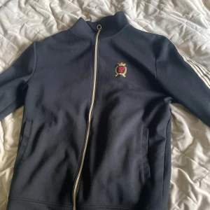 Svart ziptröja från SikSilk med brodyr - Svart ziptröja från SikSilk med broderad logga på bröstet och stora broderade bokstäver på ryggen. Tröjan har hel dragkedja, ståkrage och vita ränder längs ärmarna. Perfekt för en sportig och trendig look.