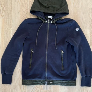 Moncler Cardigan - Snygg mörkblå cardigan/Ziphoodie från Moncler med huva i olivgrönt och matchande detaljer vid fickor och nederkant. Jackan har dragkedja framtill, två sidofickor med dragkedja och ribbade muddar. Insidan är mjukt fodrad för extra komfort. Perfekt för dig som gillar stilrena och sportiga plagg. Size S, TTS. Avtagbar luva om man vill. Fint skick! Fler frågor tas i pm!/Neo💫