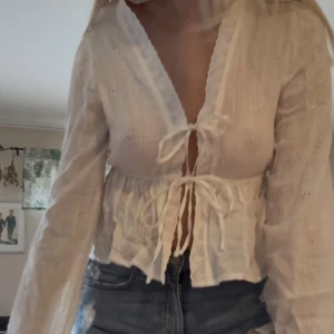 Vit blus med knyt och spetsdetaljer - Säljer en söt vit blus med v-ringning, knyt framtill och spetskant längs urringningen. Blusen har långa ärmar, broderade små hålmönster och volang nedtill. Perfekt till jeans för en romantisk look.