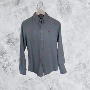 Blå skjorta från Polo Ralph Lauren - Klassisk ljusblå skjorta från Polo Ralph Lauren i custom fit. Skjortan har button-down krage, knappar framtill och broderad logga på bröstet. Perfekt för en clean och stilren look.