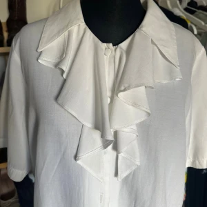 Vit volangskjorta från Lindex Connect - Säljer en vit skjorta från Lindex Connect med stora volanger framtill och korta ärmar. Skjortan har klassisk krage och knäppning framtill. Perfekt för dig som vill sticka ut med en unik och trendig look.