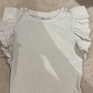 Vit ribbad topp med volangärmar Zara - Supersöt vit ribbad topp från Zara med volangdetaljer på axlarna. Toppen är ärmlös och har en rund halsringning. Perfekt för dig som gillar en feminin och trendig stil. Materialet är mjukt och stretchigt för extra komfort.
