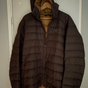 Brun  dunjacka från Arc'teryx - Säljer en svart dunjacka från Arc'teryx med huva och dragkedja framtill. Jackan är quiltad och har en lätt oversized passform, perfekt för kalla dagar. Klassisk design med diskret logga på bröstet och praktiska sidofickor.