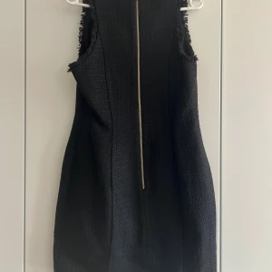 Svart ärmlös kortklänning med guldknappar - Snygg svart ärmlös kortklänning med markerad v-ringning och sex dekorativa guldknappar framtill. Klänningen har råa kanter längs ärmhål och sidor, samt en synlig dragkedja i ryggen. Materialet är strukturerat och ger en exklusiv känsla.