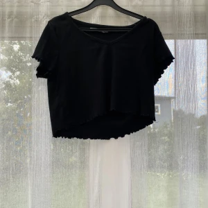 Svart croppad ribbad topp 157 - Svart croppad topp från 157 med ribbad struktur och v-ringning. Toppen har korta ärmar och vågig kant nertill och vid ärmslut. Perfekt för dig som gillar en enkel men trendig stil.