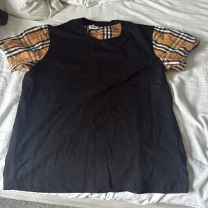 Svart burrberry t-shirt - Svart t-shirt med klassiska rutiga korta ärmar i beige, svart, vitt och rött. T-shirten har rund hals och är i mjuk bomull. Perfekt för dig som vill ha en basic tee med en twist och snygga detaljer på ärmarna.