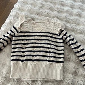 Randig stickad tröja Holly & Whyte XS - Supersöt stickad tröja från Holly & Whyte i beige och marinblått med kabelstickade detaljer, volang på axlarna och rund hals. Tröjan har lång ärm och ett klassiskt randigt mönster som ger en preppy vibe.