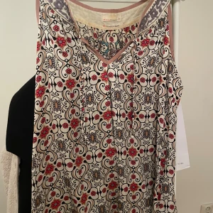 Odd Molly blommig ärmlös blus - Supersnygg ärmlös blus från Odd Molly med blommigt mönster i rött, beige och svart på vit botten. V-ringad med ljusrosa kant och broderad detalj i nacken. Luftig och flowy modell, perfekt för varma dagar.