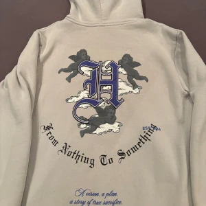 Beige hoodie från Hoodrich med tryck - Säljer en beige hoodie från Hoodrich med stora tryck på både fram- och baksida. Motiven är svarta änglar, moln och gotisk text. Hoodien har huva, långa ärmar och är i mjuk bomull. Näst intill oanvänd vädligt unik design. Passar både S och M       .     🚨OBS⚠️ du får byxorna på köpet!