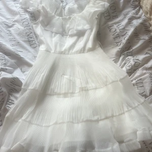 Vit plisserad kortklänning med volang - Superfin vit kortklänning med plisserad kjol och volangdetaljer upptill. Klänningen har v-ringning och är ärmlös, med ett medföljande bälte i midjan för extra stil. Perfekt för sommarens alla tillfällen och ger en fräsch och luftig känsla. Aldrig använd då jag inte kunde lämna tillbaka. Från miss rosier.