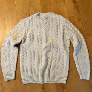 Kabelstickad Dobber - Säljer en ljus, kabelstickad tröja från Dobber i storlek medium. Tröjan har klassiskt flätmönster, rund halsringning och ribbade muddar vid ärmslut och nederkant. Perfekt för kyliga dagar och enkel att matcha med jeans eller chinos.