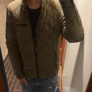 Olivgrön quiltad jacka från Replay - Snygg olivgrön quiltad jacka från Replay med dragkedja framtill och svarta detaljer. Jackan har lång ärm, ståkrage och flera praktiska fickor med dragkedja. Perfekt för dig som vill ha en stilren och bekväm jacka med modern känsla. Jackan har storlek s och modellen är 173 cm!