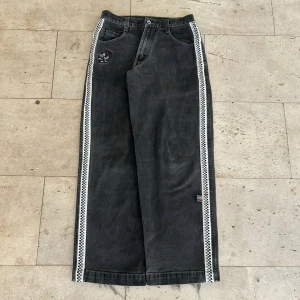 Svarta wide jeans med checkerband - Unika svarta jeans med bred passform och checkerband längs sidorna. Byxorna har broderad patch med tecknad fisk vid fickan och coola detaljer som ger streetkänsla. Tillverkade i jeansmaterial och med raka ben för avslappnad vibe.