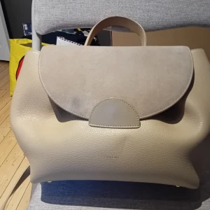 Polène Paris Un väska  - Snygg och stilren handväska från Polène Paris i beige skinn och mocka. Väskan har ett rundat lock, handtag upptill och invändig ficka med dragkedja. Perfekt för dig som gillar minimalistisk design och neutrala färger.