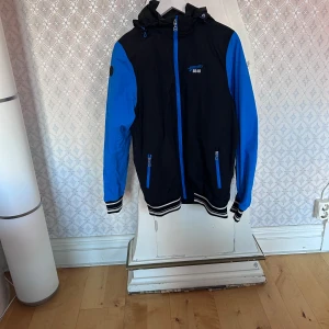 Svart och blå vindjacka 8848 Altitude - Snygg svart och blå vindjacka från 8848 Altitude med huva, meshfoder och dragkedjor i blått. Jackan har ribbade muddar med vita ränder och logga på bröstet samt ärmen. Perfekt för dig som gillar sportig stil och vill sticka ut.