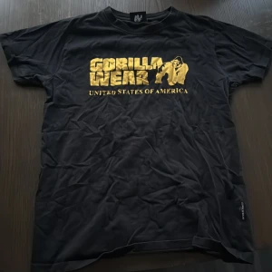 Svart Gorilla Wear t-shirt i guldtryck - Svart t-shirt från Gorilla Wear med stort guldtryck på bröstet och texten 'United States of America'. Klassisk passform och rund hals. Perfekt för dig som gillar sportig och street-inspirerad stil.