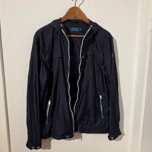 Svart vindjacka från Polo Ralph Lauren - Svart vindjacka från Polo Ralph Lauren med huva och vita dragkedjor. Jackan har två fickor med dragkedja framtill och diskret logga på ärmen. Lätt och smidig modell i syntetmaterial, perfekt för blåsiga dagar.