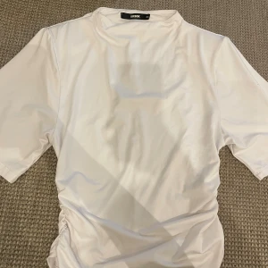 Vit t-shirt med snörning från Bikbok - Vit t-shirt från Bikbok i storlek XS. Modellen har korta ärmar och cool snörningsdetalj på sidan som ger en snygg twist. Den har också axelvaddar. Materialet känns mjukt och stretchigt, perfekt för dig som gillar enkel men trendig stil.