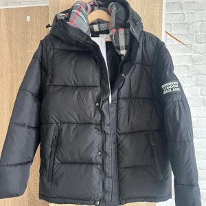 Svart pufferjacka från Burberry - Säljer en svart pufferjacka från Burberry med huva och dragkedja. Jackan har ett klassiskt rutigt foder i grått, vitt och rött samt logga på ärmen. Yttermaterialet är polyester och fodret är mjukt och varmt. Perfekt för kyliga dagar.