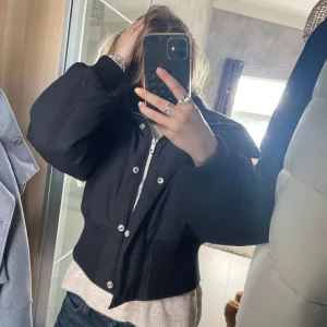 Svart pufferjacka med ribbade detaljer - Säljer en svart pufferjacka med bred ribbad krage och muddar. Jackan är croppad och har både dragkedja och knappar framtill. Perfekt för dig som vill ha en cool och oversized look. Passar till jeans och sneakers för en chill vibe.