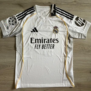 Real Madrid Bellingham hemma matchtröja M - Säljer en officiell Real Madrid fotbollströja från Adidas i storlek M, med Bellingham och nummer 5 på ryggen. Tröjan är vit med svarta detaljer, gula inslag längs sidorna och klubbmärke på bröstet. Tillverkad i återvunnet material och har Champions League-märke på ärmen.
