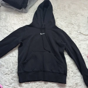 Svart Nike hoodie XS - Svart hoodie från Nike i storlek XS med klassisk vit swoosh-logga på bröstet. Tröjan har huva med snörning och en stor känguruficka framtill. Perfekt för en chill och sportig look. Tillverkad i mjuk bomullsblandning för extra komfort. Använt väldigt få gånger då den är för kort för mig, nypris 800kr.
