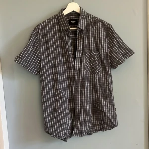 Grå kortärmad skjorta  - Rutig gråvit kortärmad skjorta. Spänne på ärmarna. Storlek M. Gott skick, inga defekter eller missfärgningar.