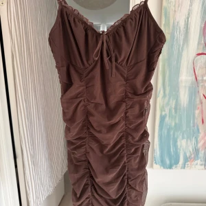 Brun meshklänning med rynk  - Säljer en brun kortklänning från H&M i mesh med smala axelband, rynkade detaljer framtill och söt knytning vid urringningen. Klänningen har en figurnära passform 