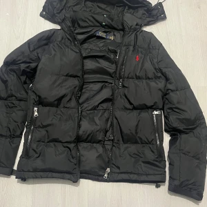 Polo Ralph Lauren jacka - Svart pufferjacka från Polo Ralph Lauren med huva och dragkedja framtill. Jackan har två stora fickor med dragkedja och en röd broderad logga på bröstet. Perfekt för kalla dagar och har en klassisk, sportig look. M på storleken men rekommenderar den för dem som har S 