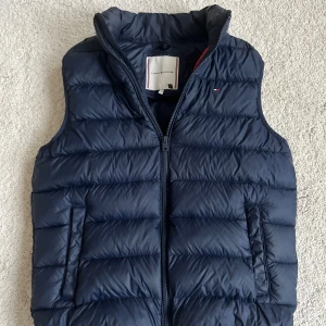  Tommy hilfiger väst - Snygg mörkblå dunväst från Tommy Hilfiger med quiltad design och dragkedja framtill. Västen har två fickor på sidorna och klassisk ståkrage. Insidan har röda och vita detaljer som ger en cool kontrast. Perfekt för lager-på-lager-stil.