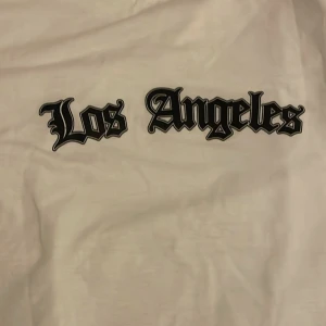 Vit Los Angeles t-shirt i bomull - Vit t-shirt med trycket 'Los Angeles' i svart gotisk stil på bröstet. Klassisk rund hals och korta ärmar, tillverkad i mjuk bomull. Perfekt för dig som gillar streetwear och vill ha en clean look med coolt tryck.