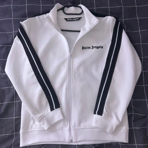 Vit track jacket från Palm Angels - Säljer en stilren vit track jacket från Palm Angels med svarta ränder längs ärmarna. Jackan har dragkedja framtill och en broderad logga på bröstet. Perfekt för en sportig look!
