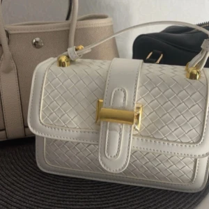 Vit axelväska med guldiga detaljer - Snygg vit axelväska i flätad skinnimitation med guldiga metalldetaljer och spänne framtill. Väskan har ett stilrent och lyxigt utseende, innerfack med dragkedja och justerbart axelband. Perfekt för dig som gillar trendiga accessoarer.