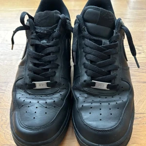 Svarta Nike Air Force sneakers - Säljer ett par svarta Nike Air Force sneakers i bra skick. De är tillverkade i skinn och har klassisk snörning. Perfekta för både vardag och fest. Storlek är inte angiven, men de ser ut att passa en normal passform. Skorna har en stilren design med perforerade detaljer på ovansidan.