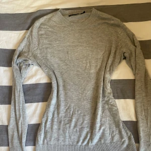 Grå stickad tröja med rund hals - En enkel och stilren grå stickad tröja med rund halsringning och långa ärmar. Tröjan har ribbade muddar vid ärmslut och nederkant, perfekt för lager-på-lager under kyligare dagar.