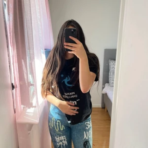 Blå baggy jeans med graffiti-tryck - Säljer ett par blå baggy jeans med coola graffiti-inspirerade tryck och textdetaljer i vitt, gult och svart. Byxorna har hög midja och raka ben med avslappnad passform. Perfekta för dig som vill sticka ut med en unik streetstyle-look.