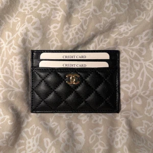 Svart quiltad korthållare - Supersnygg svart korthållare i klassiskt äkta skinn med guldfärgad logga framtill. Smidig och stilren design med flera kortfack både fram och bak. Perfekt accessoar för dig som vill ha lyxig känsla i vardagen.