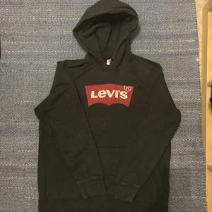 Svart hoodie från Levi's med logga - Svart hoodie från Levi's i storlek L med den klassiska röda och vita Levi's-loggan på bröstet. Tröjan har huva, lång ärm och är tillverkad i 100% bomull för skön komfort. Perfekt för dig som gillar streetwear och vill ha en enkel men snygg look.