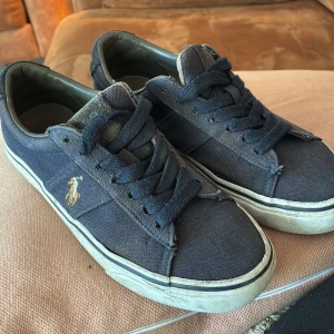 Mörkblå sneakers från Polo Ralph Lauren - Snygga mörkblå sneakers från Polo Ralph Lauren med klassisk logga broderad i vitt och guld på sidan. Skorna har snörning, platt sula och är tillverkade i canvas med mockadetalj på hälen. Perfekta för en avslappnad och stilren look.
